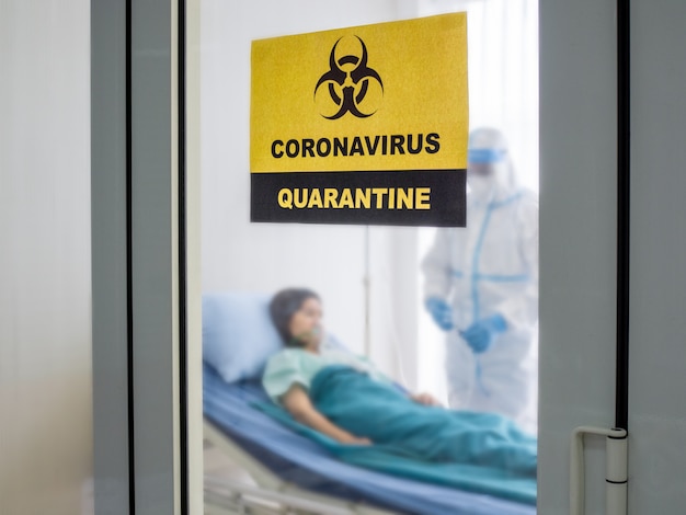 Asiatischer Arzt trägt einen PSA-Anzug mit N95-Maske und Gesichtsschutz, behandelt mit Coronavirus infizierten Patienten im Unterdruckraum und ist mit einem Schild mit Quarantänealarmbereich gekennzeichnet.