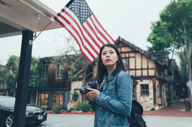 asiatischer Backpacker-Fotograf, der in den Frühjahrsferien auf Straßenfotos im urbanen Stil fotografiert. Mädchen Reisende mit professioneller Kamera auf dem Dach mit amerikanischer Flagge in Carmel auf dem Seeweg