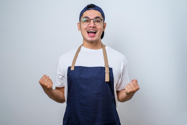 Asiatischer barista-mann mit schürze, der sehr glücklich und aufgeregt steht und eine siegergeste mit erhobenen armen macht, lächelt und schreit nach erfolg und blickt in die kamera feierkonzept