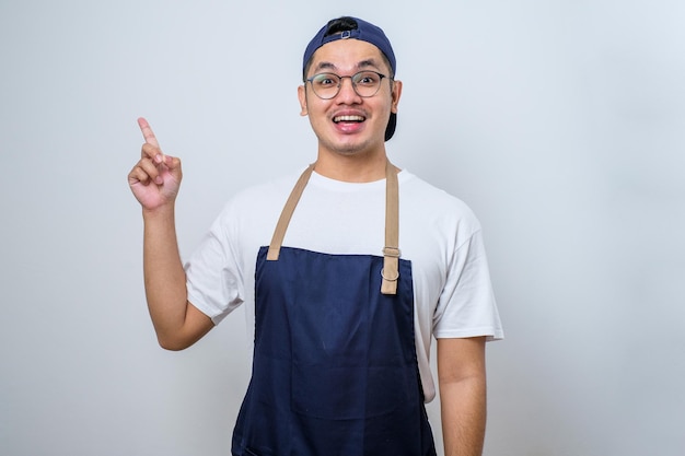 Asiatischer Barista-Mann mit Schürze und Mütze, der mit den Fingern Nummer eins nach oben zeigt und dabei selbstbewusst und glücklich lächelt