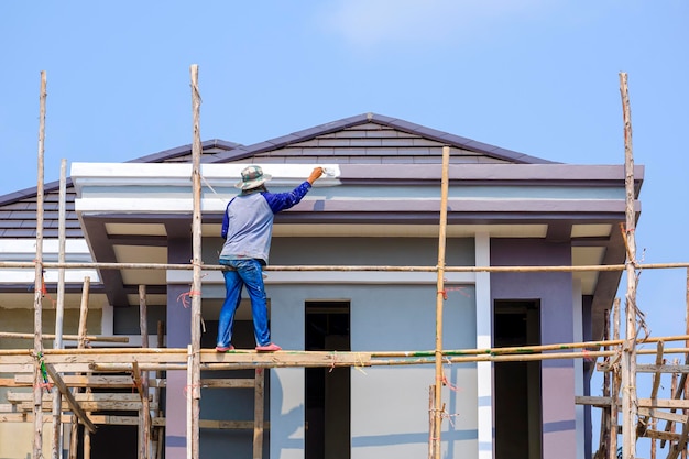 Asiatischer Bauarbeiter auf Holzgerüst malt Dachkonstruktion eines modernen Hauses