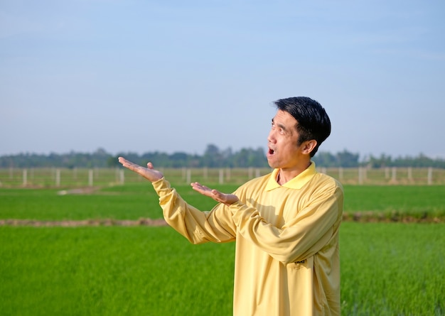 Asiatischer Bauer trägt ein gelbes Hemd im Stehen und hebt eine Hand mit einem Wow-Überraschungsgesicht auf der grünen Farm?