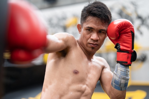 Asiatischer Boxersportler mit roten Boxhandschuhen trainiert, indem er im Fitnessstudio gegen Sandsäcke schlägt