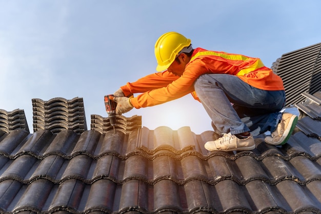 Asiatischer Dachdecker, der an der Dachkonstruktion des Gebäudes auf der Baustelle arbeitetRoofer mit Luft- oder pneumatischer Nagelpistole und Installation von Betondachziegeln auf dem neuen Dach