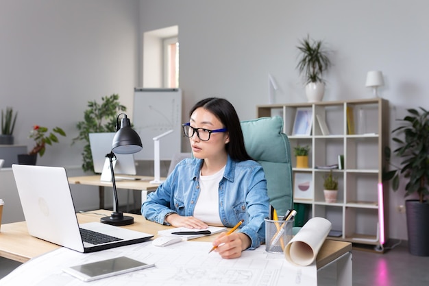 Asiatischer Designer, der im Büro mit Blaupausen arbeitet Frau bei der Arbeit in Freizeitkleidung