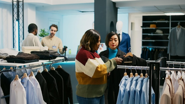 Asiatischer Einzelhändler, der einem Kunden bei der Auswahl von Kleidung hilft, verwendet ein Tablet, um den Warenbestand im Kaufhaus zu überprüfen.