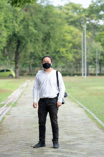 Asiatischer freiberuflicher Langhaarmann mit schwarzer Gesichtsmaske steht und postet mit seiner Kamera neben ihm auf dem Gehweg im Garten im Freien