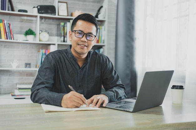 Asiatischer freiberuflicher Mann mit Brille und Lächeln, der zu Hause auf dem Laptop tippt und Notizen in das Notizbuch schreibt Unternehmermann, der für Unternehmen im Wohnzimmer zu Hause arbeitet Geschäftskonzept für die Arbeit zu Hause