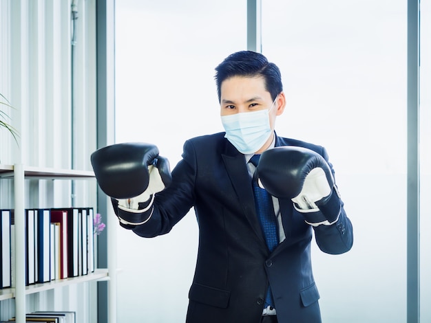 Asiatischer Geschäftsmann, der Anzug und schützende Gesichtsmaske und Hand trägt, die Boxhandschuhe im Büro tragen