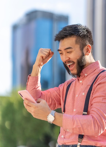 Asiatischer Geschäftsmann, der Erfolg gestikuliert, während er das Handy im Freien verwendet