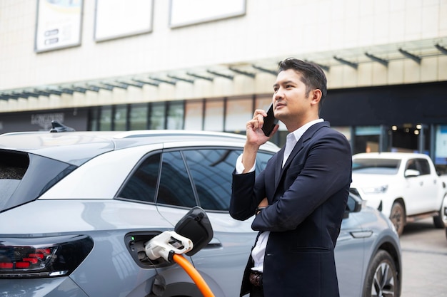 Asiatischer Geschäftsmann, der mit einem Elektroauto unterwegs ist und an einer Ladestation anhält, die mit einem Internetkabel auf dem Smartphone verbunden ist und freudig lächelt, während er die energiesparende Ansicht des Elektroautos auflädt