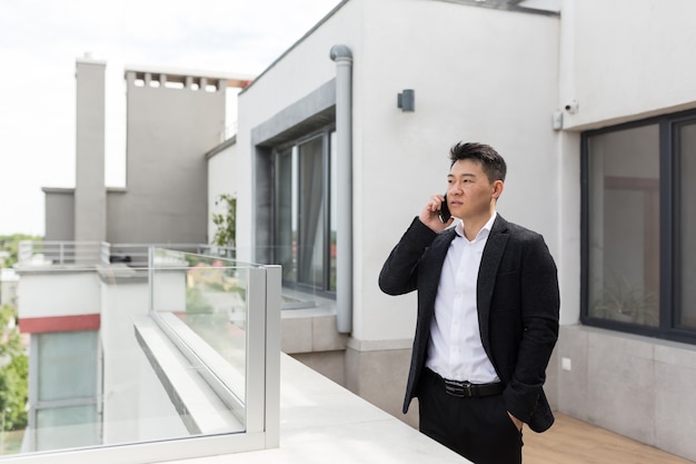 Asiatischer Geschäftsmann im Anzug genießt das Telefon auf der Balkonterrasse des modernen Bürozentrums