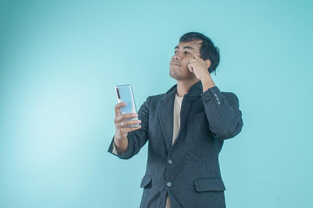 Asiatischer Geschäftsmann im schwarzen Anzug sieht nachdenklich aus, während er das Smartphone in der Hand hält. Stehen auf einem Blau