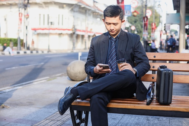 Asiatischer Geschäftsmann in einem formellen Geschäftsanzug, der auf einem Handy schaut und draußen auf einer Stadtstraße sitzt