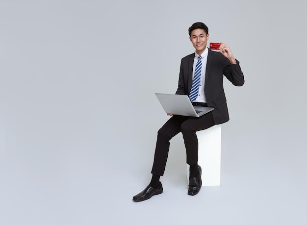 Asiatischer Geschäftsmann lächelt im formellen Anzug, der mit Laptop auf Stuhl sitzt und Kreditkarte zeigt