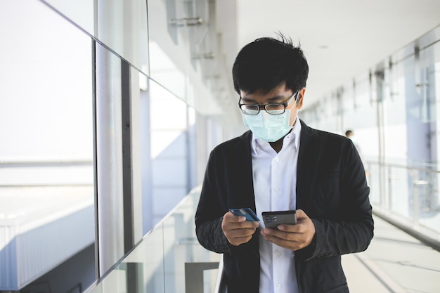 asiatischer geschäftsmann mit brille und chirurgischer maske, der online-dienste mit kreditkarte bezahlt