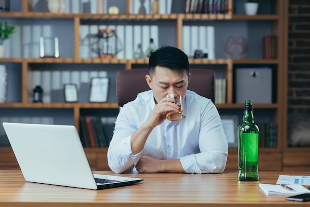 Asiatischer Geschäftsmann scheitert Mann in Verzweiflung trinkt Schnaps, der spät am Tisch im Büro sitzt, deprimiert und hoffnungslos