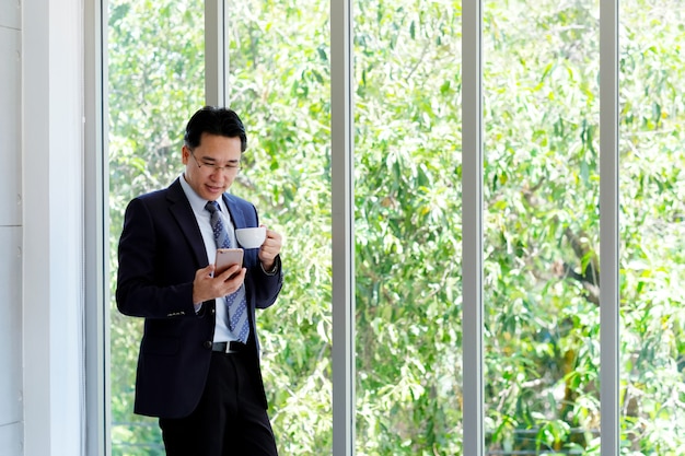 Asiatischer Geschäftsmann unter Verwendung des intelligenten Telefons und des trinkenden Kaffees im Büro, im Büroleben und im Geschäftskonzept