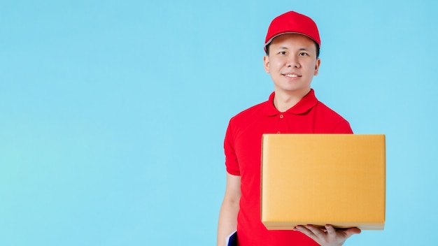 Asiatischer glücklicher Lieferer, der ein rotes Hemd trägt, das beim Halten von Papierpaketkästen lokalisiert auf blauem Farbhintergrund mit Kopienraum ein rotes Hemd trägt. Konzept des Postzustelldienstes.