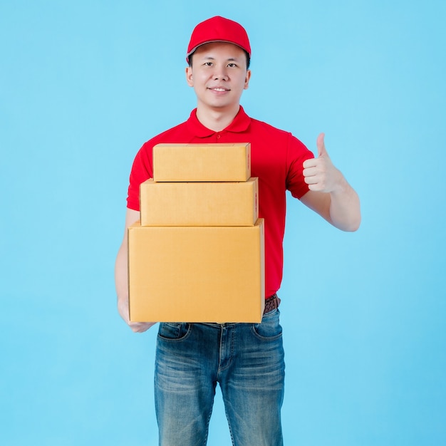 Asiatischer glücklicher Liefermann, der ein rotes Hemd und einen Hut trägt, der Daumen hoch zeigt und Papierpaketkästen einzeln auf blauem Farbhintergrund trägt. Konzept des Postzustelldienstes.