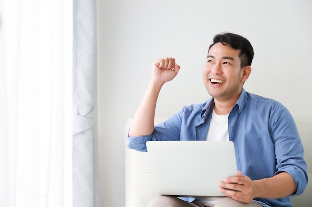 Asiatischer gutaussehender Mann, der mit Laptop-Computer im Wohnzimmer glücklich und im Lächelngesicht arbeitet