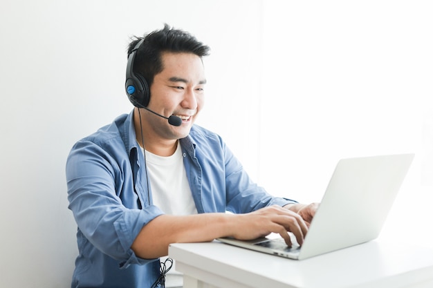 Asiatischer gutaussehender Mann im blauen Hemd unter Verwendung des Laptops mit Unterhaltungslächeln des Kopfhörers und glücklichem Gesicht