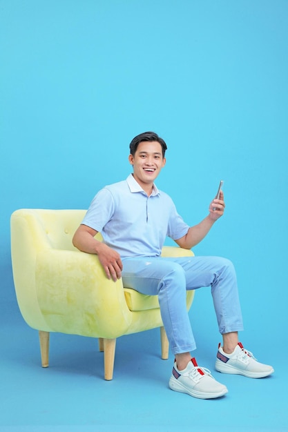 Asiatischer, gutaussehender und fröhlicher junger Mann, der ein Smartphone verwendet, sitzt auf einem Sofa, das auf dem Hintergrund isoliert ist