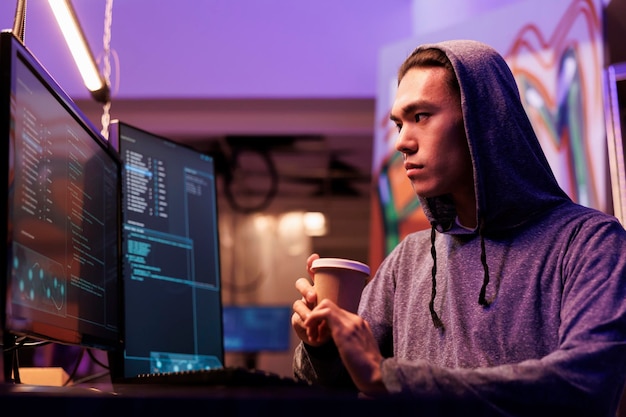 Asiatischer Hacker trinkt Kaffee und führt DDoS-Angriff auf Website durch. Krimineller bricht auf Computerbildschirm in Netzwerksystem ein, während er Getränke zum Mitnehmen in einem verlassenen Lagerhaus bereithält