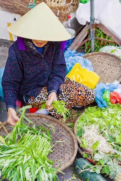 Asiatischer Händler in einem traditionellen vietnamesischen Hut, der frisches grünes Gartenmaterial auf dem Straßenmarkt in Hoi An, Vietnam, verkauft. Kein Gesicht. Selektiver Fokus