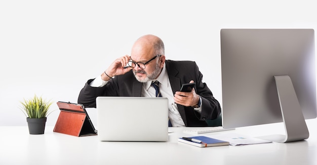 Asiatischer indischer leitender Finanzgeschäftsmann, der an seinem Arbeitsplatz oder Schreibtisch vor einem Computer, Laptop und Tablet sitzt. Telefonieren, während ich Papierkram erledige