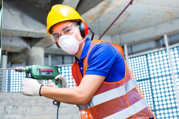 Asiatischer indonesischer Baumeister oder Arbeiter auf der Baustelle, der mit einer Maschine oder einem Bohrer, einem Gehörschutz, einer Maske und einem Helm oder Helm in einer Wand eines Turmgebäudes bohrt