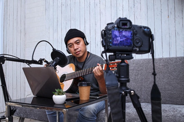 Asiatischer Influencer spielt Gitarre während Podcast oder Live-Videoübertragung für das Publikum, aufgezeichnet fr