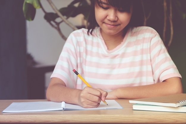 Asiatischer Jugendlich-Studentin, mit dem lächelnden Gesicht, Schreibensnotizbuchpapier auf Tabelle in ihrem Klassenzimmer, Bildungskonzept