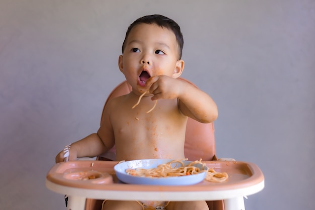 Asiatischer Junge, der auf hohem Kinderstuhl isst.