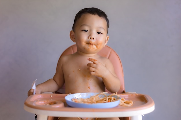 Asiatischer Junge, der auf hohem Kinderstuhl isst.