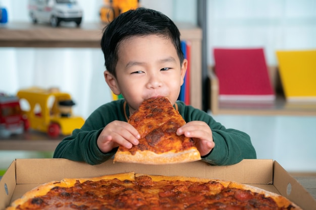 Asiatischer Junge, der Pizza und Pizzaschachtel isst, legte auf den Tisch.