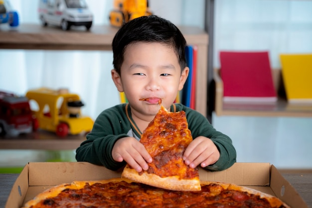 Asiatischer Junge, der Pizza und Pizzaschachtel isst, legte auf den Tisch.