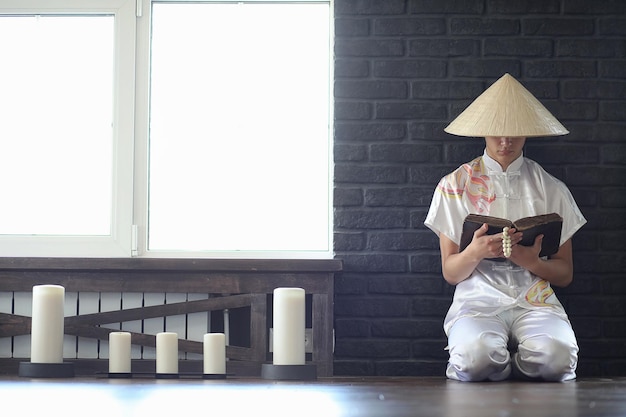 Asiatischer Junge im Kimono, der ein altes Buch liest, das am Fenster sitzt