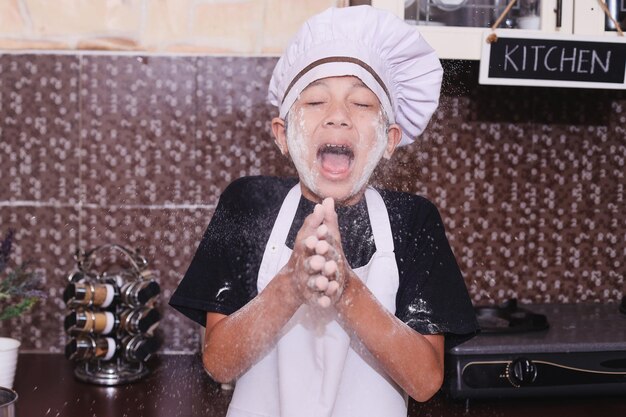 Asiatischer Junge in Kochuniform lacht und klatscht in die Hände und streut Mehl aus Teig in der Küche