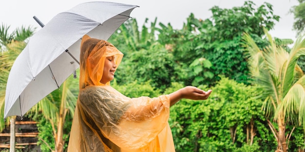 Asiatischer Junge mit orangefarbenem Regenmantel, der einen Regenschirm glücklich hält und an einem regnerischen Tag Spaß im Regen hat