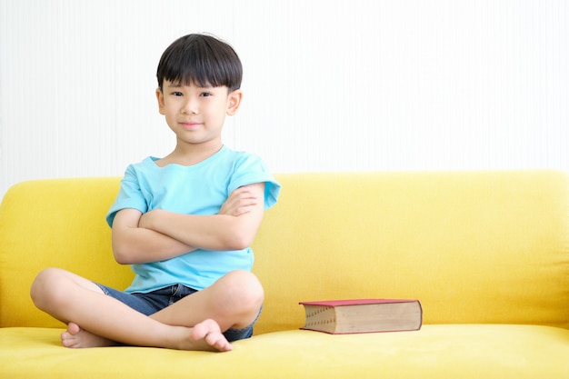 Asiatischer Junge sitzt auf gelbem Sofa mit großem rotem Buch.