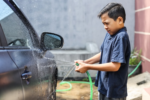 Asiatischer Junge sprüht Wasser aus dem Schlauch, um das Auto zu waschen