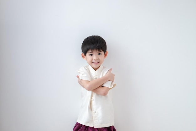 Asiatischer Junge trägt traditionelle thailändische Kleidung und steht mit verschränkten Armen auf weißem Hintergrund