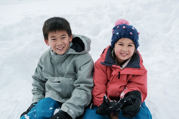 Asiatischer Junge und Mädchen der glücklichen Mischrasse, Geschwister, die auf weißem Schnee in Japan lächeln und sitzen
