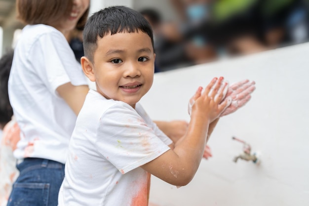 Asiatischer Junge wäscht sich die Hände mit Seife auf dem Waschbecken im Freien, nachdem er in der Schule Farbe mit seinen Händen gemalt hat