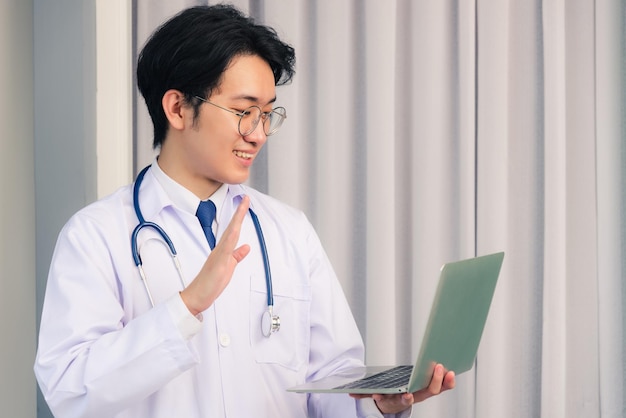 Asiatischer junger gutaussehender Arzt, der ein Arztkleid und ein Stethoskop trägt, Videokonferenz oder Facetime hebt die Hand, um Hallo zu sagen, er lächelt im Krankenhausbüro, Konzept der medizinischen Versorgung