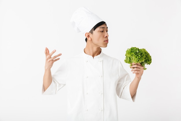 asiatischer junger Hauptmann in der weißen Kochuniform, die an der Kamera lächelt, während grüner Salat Salat gehalten über weißer Wand hält