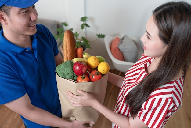 Asiatischer junger Lieferbote in der blauen Hemduniform, die Paketbeutel frisches Essen zur Frau liefert