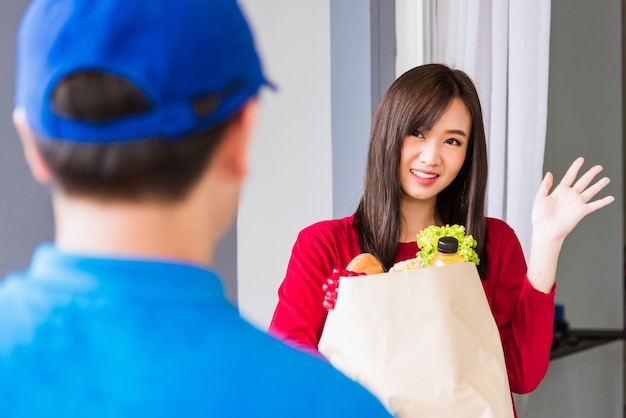 Asiatischer junger Liefermann in blauer Uniform, der einen Lebensmitteldienst macht, der der Kundin im Vorderhaus nach einer Pandemie-Coronavirus frisches Gemüse und Obst und Lebensmittel in Papiertüten gibt, Zurück zur neuen Normalität