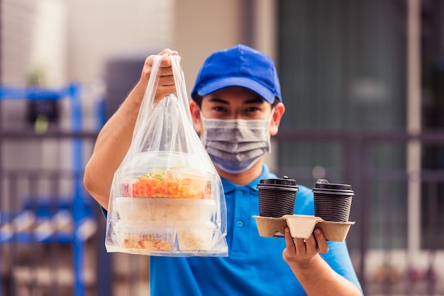 Asiatischer junger Liefermann in blauer Uniform mit Gesichtsmaske, der Lebensmitteldienst macht und Reisfutterboxen, Plastiktüten und Kaffee im Vorderhaus unter pandemischem Coronavirus gibt, Zurück zum neuen normalen Konzept